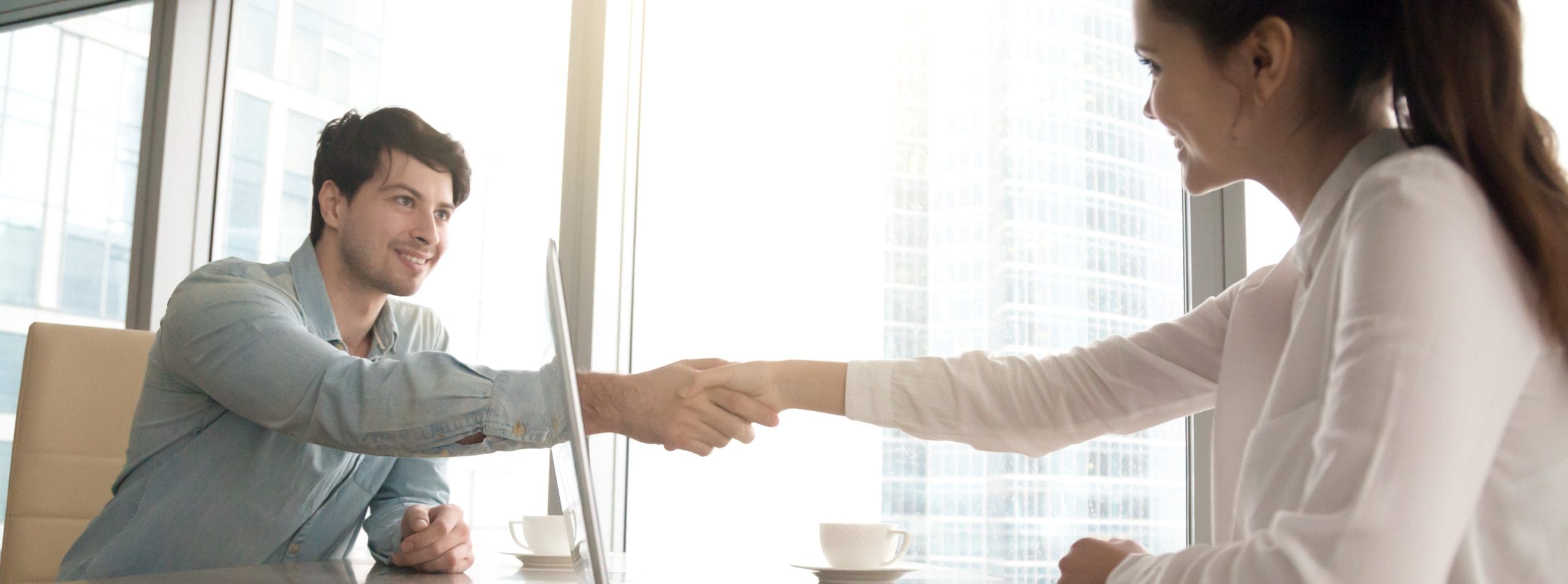 Man and woman in a bright office shaking hands across a table, with laptops and coffee cups. They appear professional and friendly.