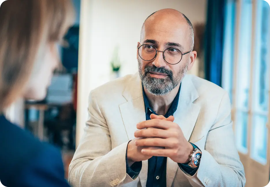 Bald man with glasses and a beard, wearing a light-colored blazer, sits across a table from another person in a dimly lit room.