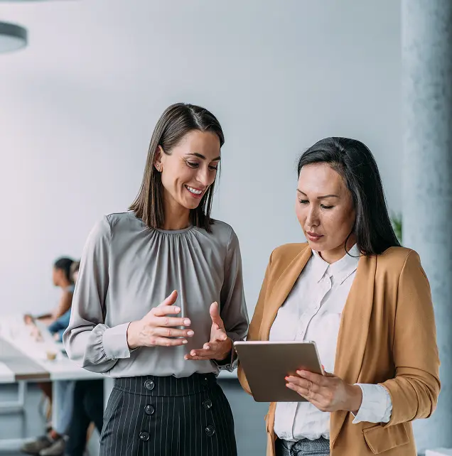 Two women engaged in a conversation while looking at a tablet in an office setting. One woman, wearing a grey blouse, appears animated and is gesturing, while the other woman, dressed in a brown blazer, is focused on the tablet.