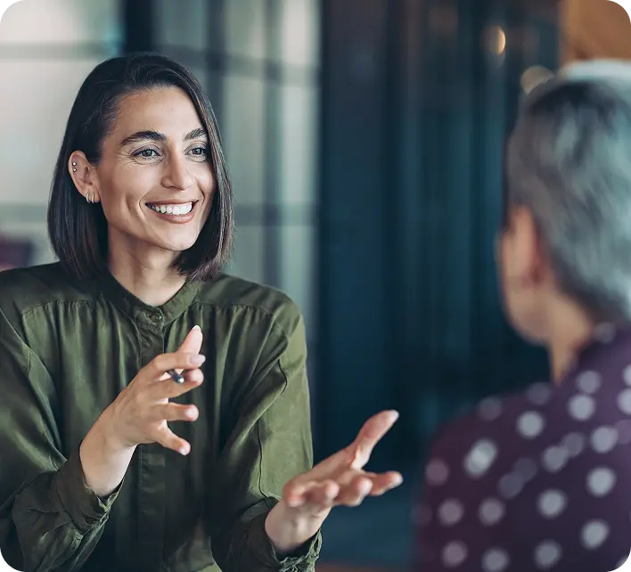 hero-solutions A woman with shoulder-length hair and wearing a green button-down shirt is smiling and gesturing as she engages in conversation with a person out of focus, seen from behind. The setting appears to be a modern, well-lit space with glass elements in the background.