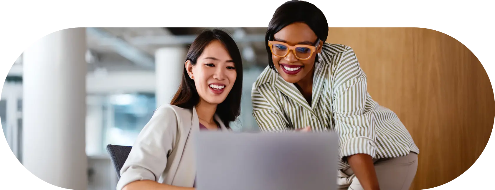 Two women smiling and looking at a laptop screen in an office setting.