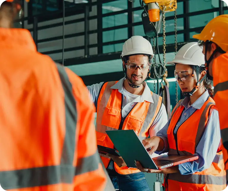 Who-we-serve-taft-hartley-funds A group of construction workers wearing orange safety vests, hard hats, and safety glasses, gathered around a laptop on a construction site. They appear to be discussing or reviewing information displayed on the laptop screen.