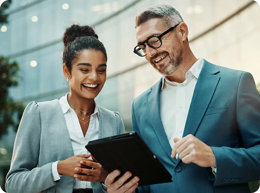 broker-hero Two business professionals in suits smiling and looking at a tablet outside a modern glass building.