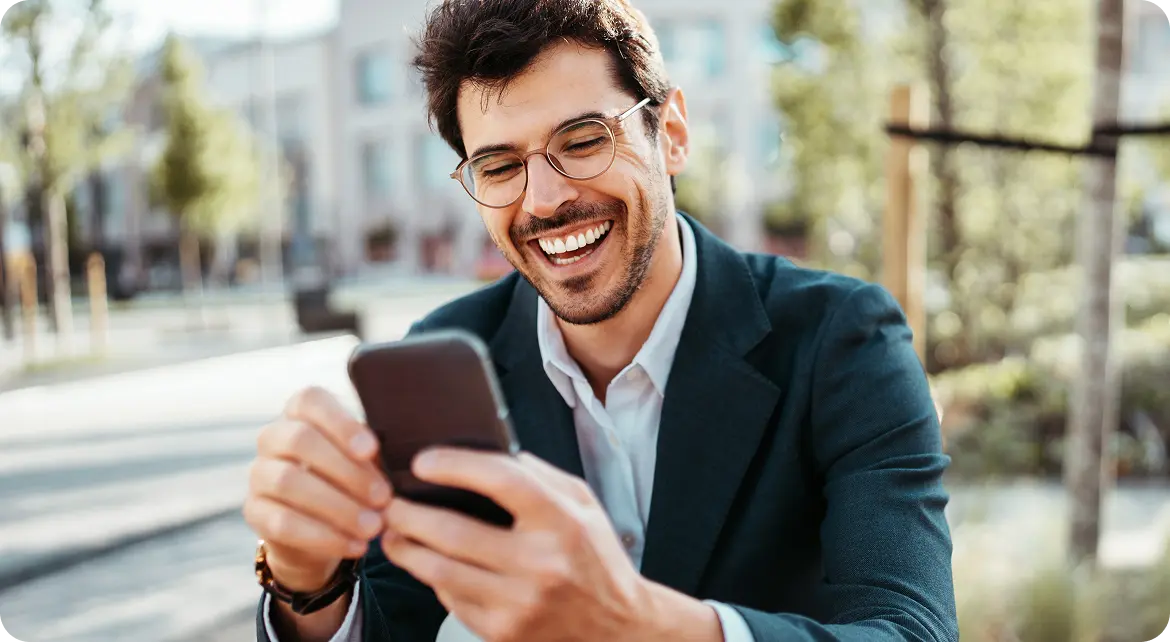 A smiling man wearing glasses and a suit is sitting outside, holding a smartphone and looking at it with amusement.