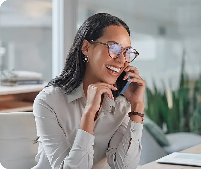 A smiling woman wearing glasses and a light-colored blouse is talking on a smartphone, sitting in a modern office environment with plants in the background.