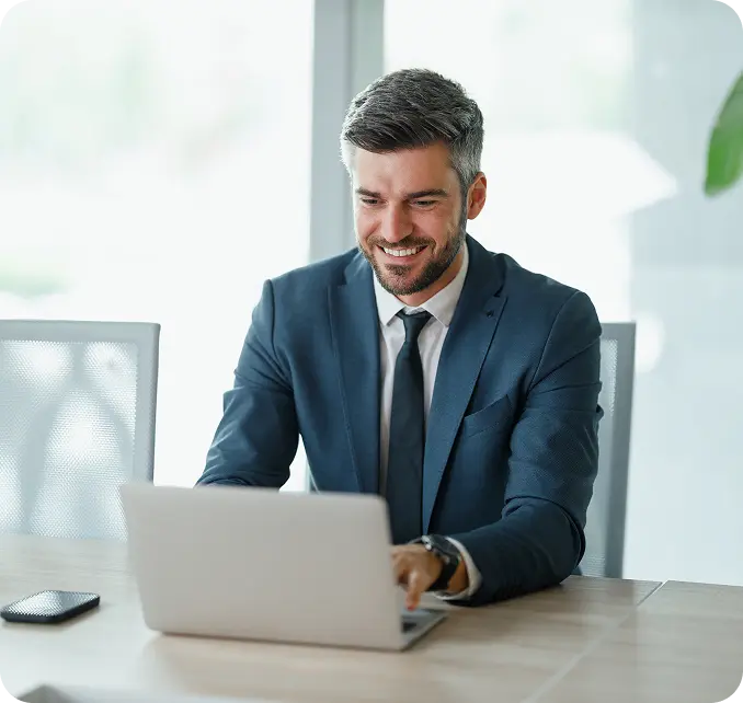 employers-hero A man in a dark suit and tie is smiling while working on a laptop at a desk, with a smartphone next to him.