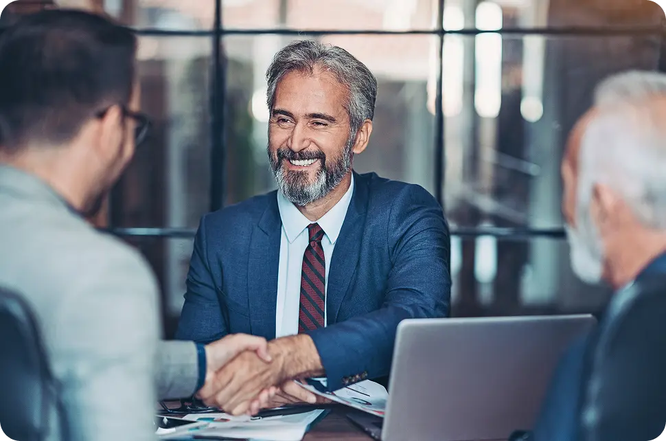 hero-abn-medical-necessity-validation A group of business professionals in suits are in a meeting room, with a smiling man with a beard shaking hands with another person across a table with a laptop and documents.