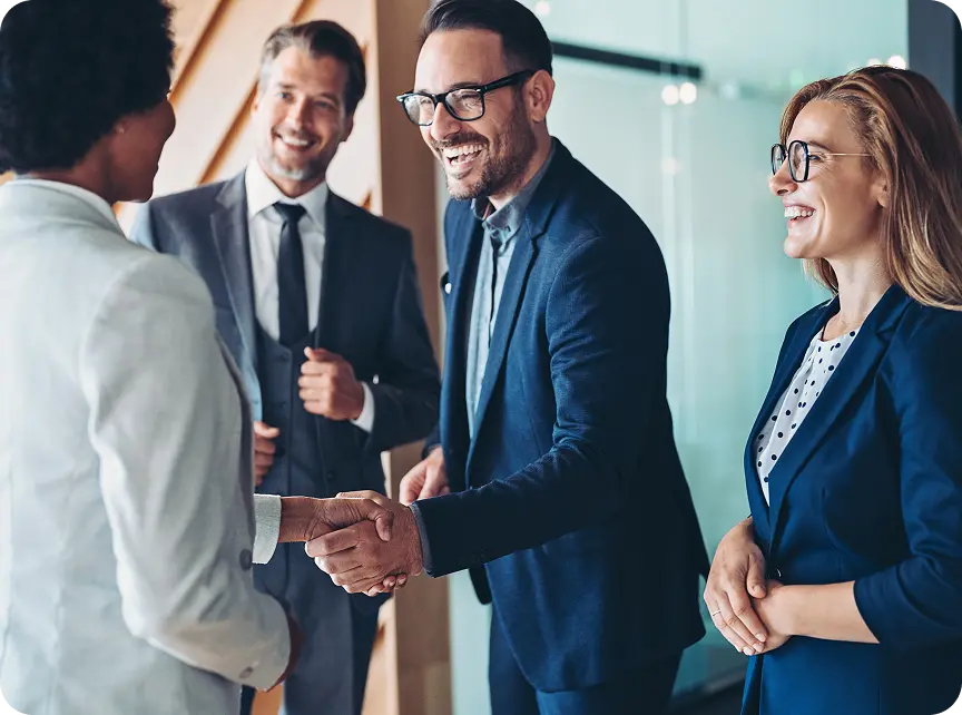 A group of four business professionals in formal attire are standing and smiling, with one man in glasses shaking hands with a woman. Two other colleagues are standing nearby, watching the interaction in a positive office environment.