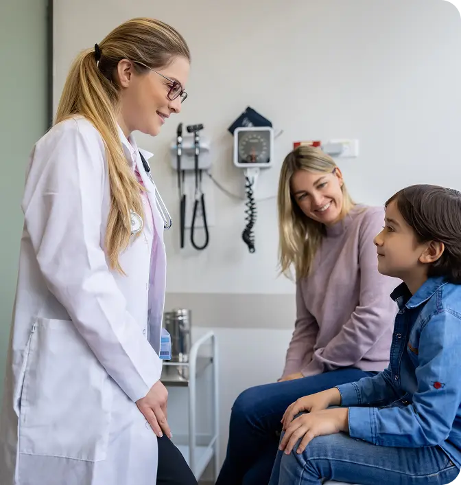 memebers-doctor-patient-examine-room A female doctor in a white coat is smiling and leaning towards a young boy sitting on an examination table, while a woman, presumably the boy's mother, sits beside him, also smiling. Medical equipment is visible on the wall in the background.