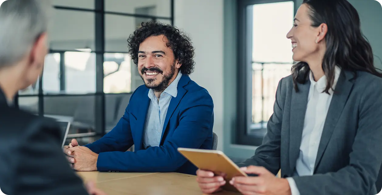 resources-overview Three professionals sitting at a conference table, engaged in a meeting, with one man in a blue suit smiling and a woman holding a tablet.