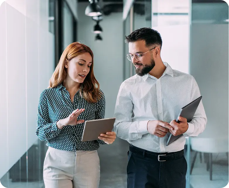 who-we-serve-facilities-providers A man and a woman walking and discussing over a tablet in an office hallway. The woman is using hand gestures while the man holds a laptop.