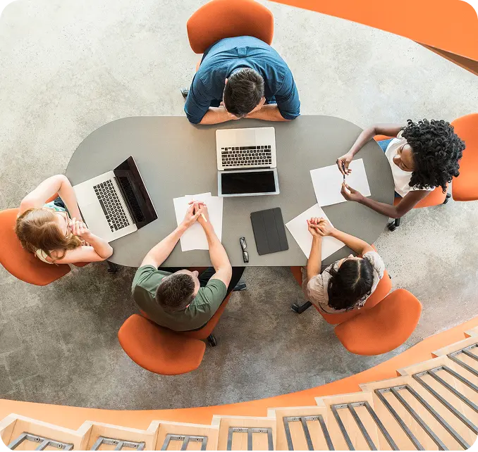 who-we-serve-health-plans Top view of six people seated around a conference table with laptops and documents, engaged in a discussion. The setting includes orange chairs and a curved staircase in the background.
