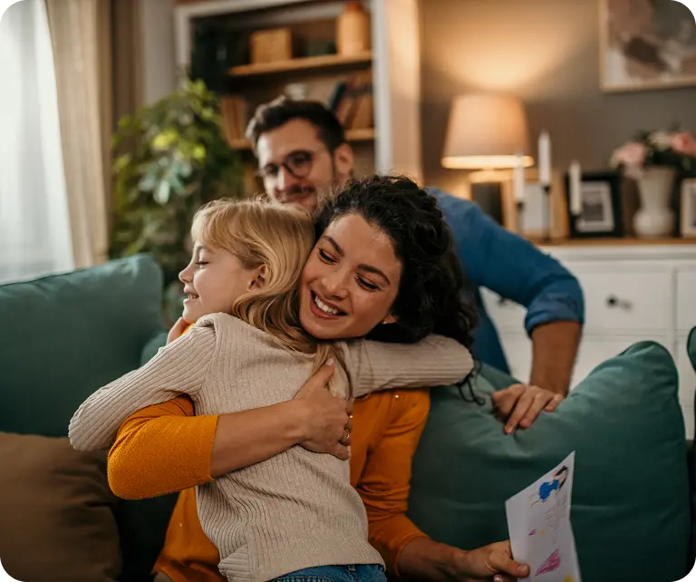 who-we-serve-members A woman and a little girl hugging each other warmly on a couch in a cozy living room, with a man smiling in the background.