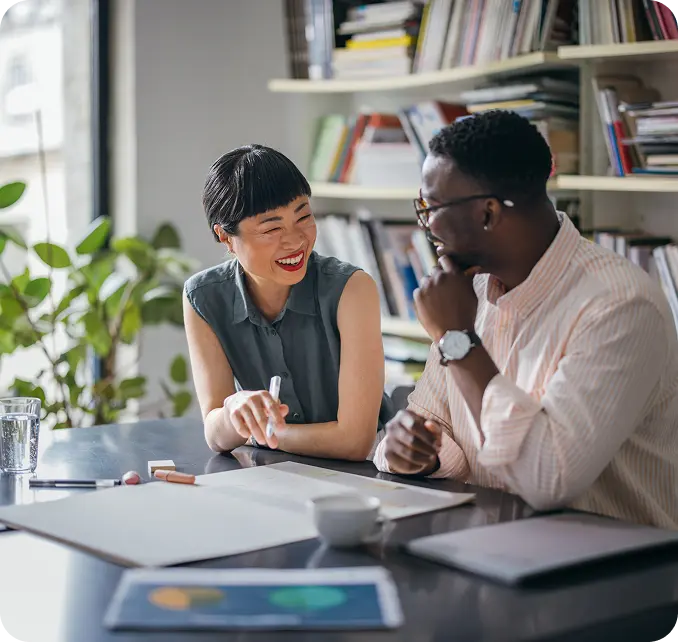 who-we-serve-stop-loss Two people are sitting at a desk in an office setting, smiling and engaged in conversation, with bookshelves and plants in the background.