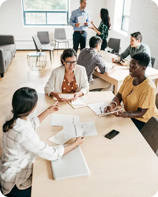 who-we-serve-tpa A group of people engaged in a meeting in a well-lit office setting, with some seated at a table discussing documents and others standing and conversing in the background, creating a collaborative atmosphere.