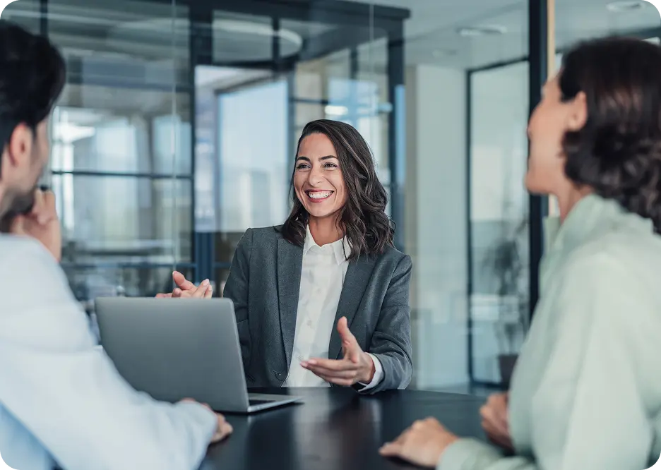 woman-2-males-business-meeting A woman in a gray blazer smiles and gestures while sitting at a conference table, engaged in a discussion with two other people. A laptop is open in front of her, and large windows provide a view of a modern office setting.