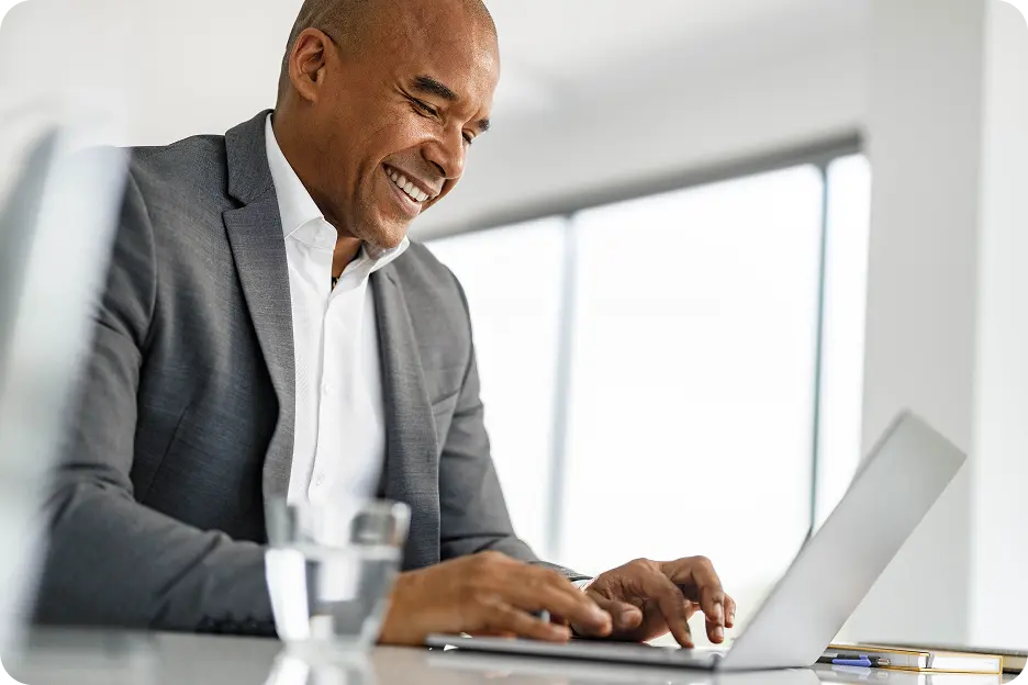 Smiling man in a grey suit working on a laptop at a bright, modern office desk with a glass of water nearby.