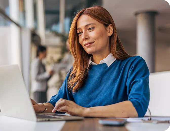 A woman with long, reddish-brown hair is sitting at a desk working on a laptop in a modern office environment, wearing a blue sweater with a white collar.