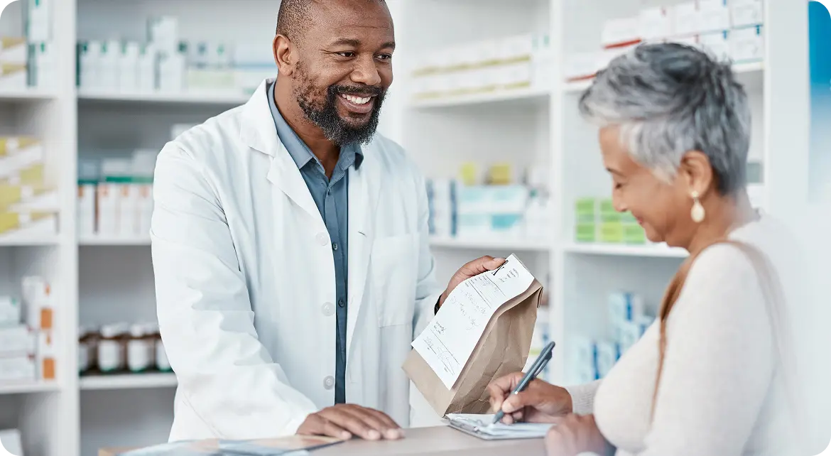 Hero-Bluebook-Rx A pharmacist in a white coat hands a paper bag to a smiling customer at a pharmacy counter, with shelves of medicines in the background.