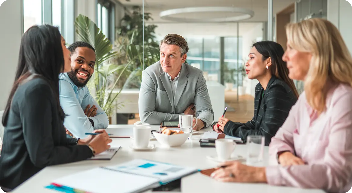 Hero-Careers A diverse group of five people sitting around a conference table, engaged in a meeting, with notebooks, coffee cups, and a bowl of snacks on the table in a bright, modern office space.