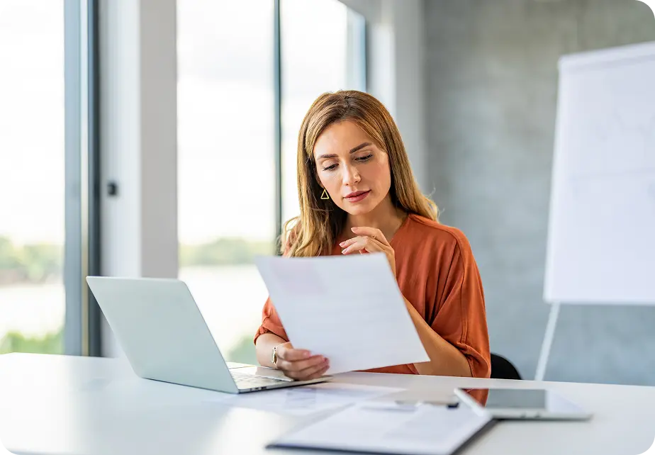 Hero-FWA A woman in an orange top sits at a desk in a modern office, reading a document with a focused expression. She is using a laptop, and there is a flipchart in the background.