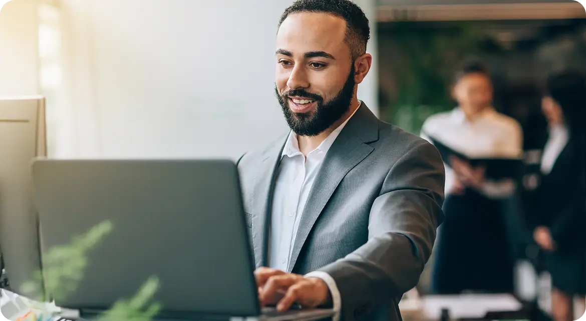 Hero-Login A man in a suit smiling while working on a laptop in an office setting, with two colleagues in the background.