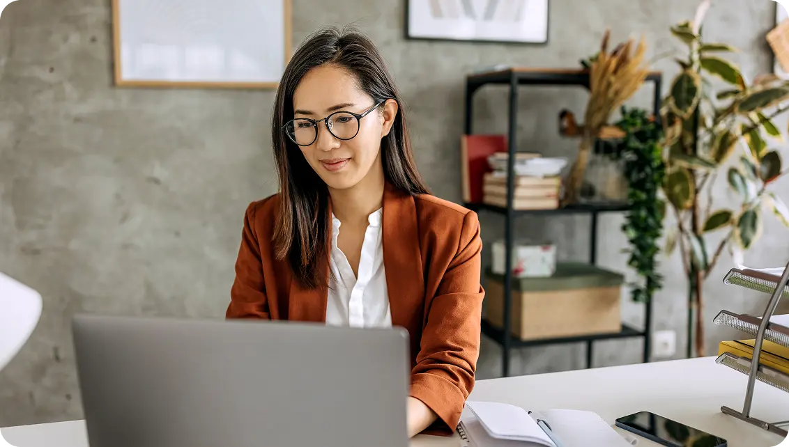 A woman wearing glasses and a brown blazer is sitting at a desk using a laptop, with a bookshelf and plants in the background.