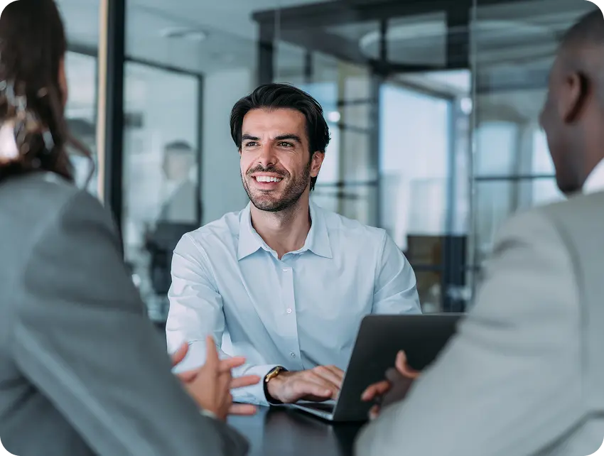 Hero-Payment-Integrity A smiling man in a light blue shirt is sitting at a conference table, engaged in conversation with two colleagues, who are partially visible. He is using a laptop, and the background features a modern office setting with glass partitions.