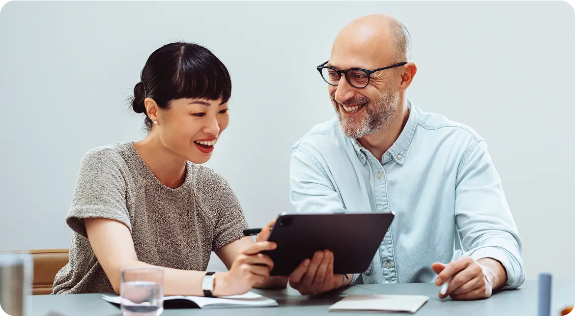 Hero-Plan-Performance A woman and a man are sitting together at a table, both smiling and looking at a tablet that the woman is holding; the man has glasses and a beard, and the setting is bright and modern.
