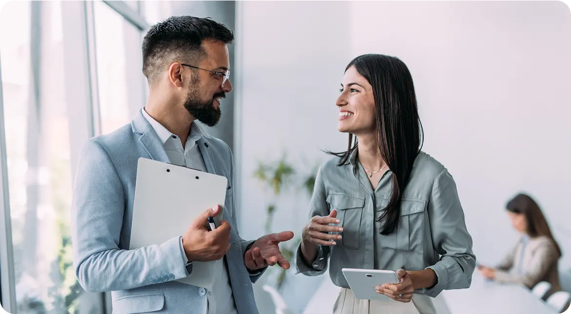 Hero-Self-Funded Two professionals smiling and conversing in a brightly lit office space, with one holding a tablet and the other holding a clipboard.