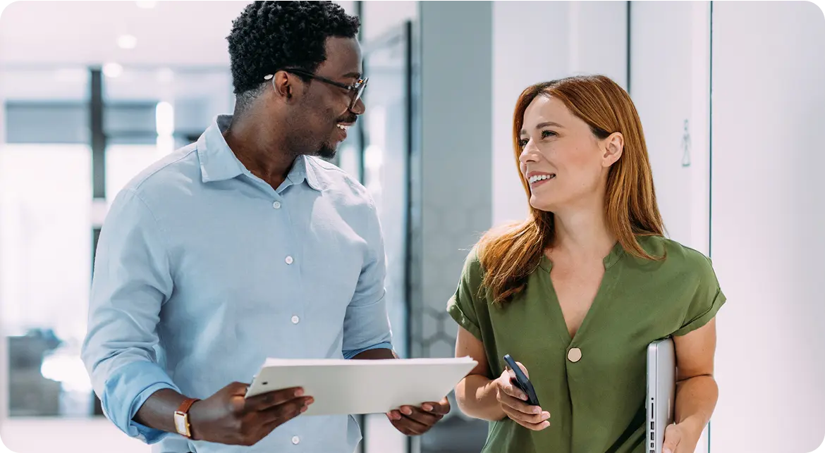 Hero-Stop-Loss Two colleagues walking down a hallway, engaging in conversation; the man on the left holds a tablet and the woman on the right carries a laptop and smartphone.