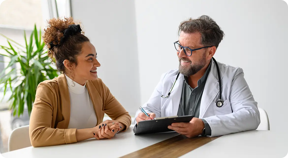 A smiling healthcare professional in a white coat is sitting across from a woman with curly hair, who is dressed in a beige cardigan. They are engaged in a friendly conversation at a table in a bright, modern office, with a plant in the background.