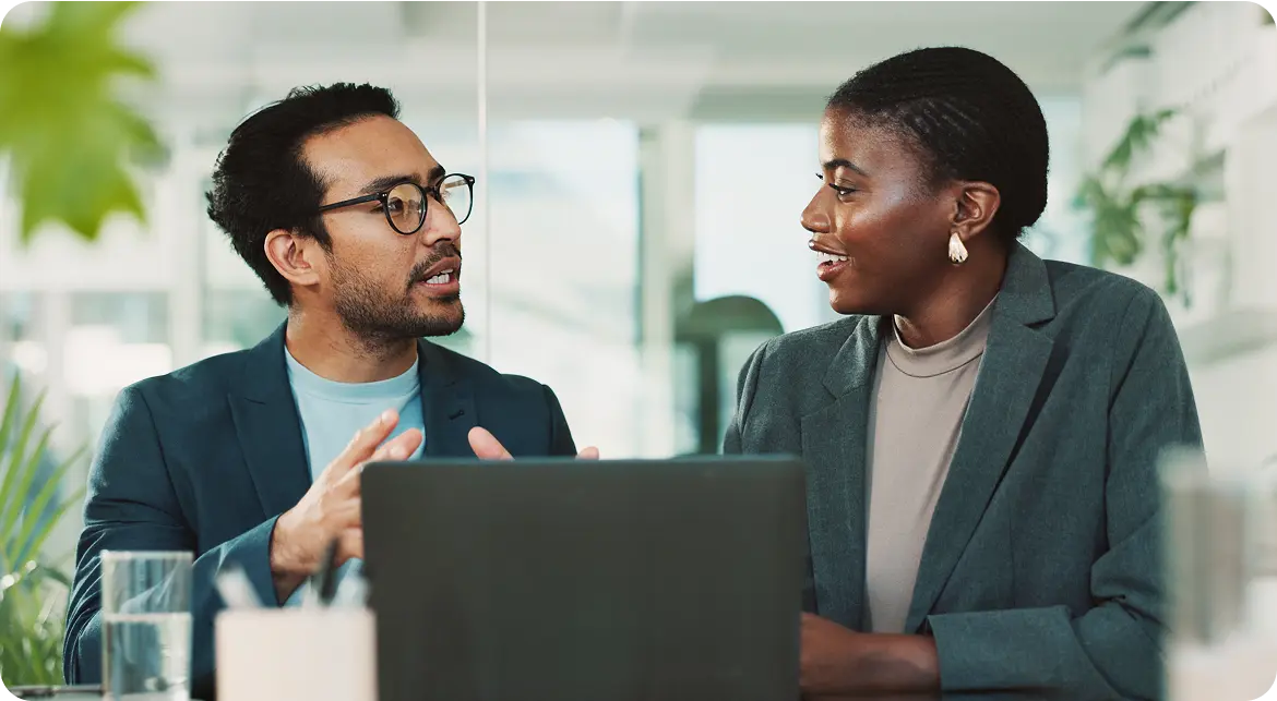 Two professionals having a discussion, seated at a table with a laptop, in a bright office setting.