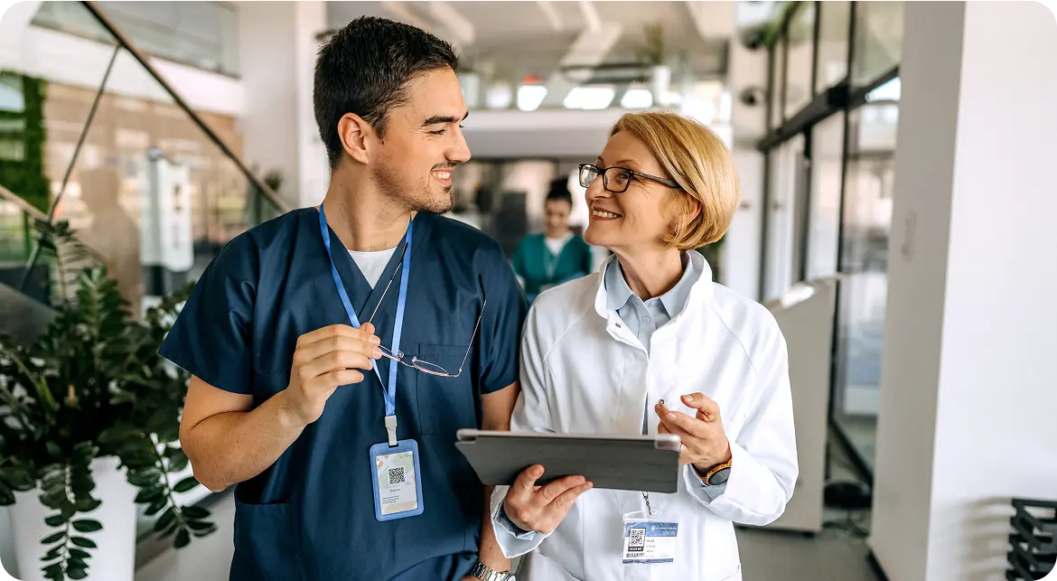 A male doctor in blue scrubs and a female doctor in a white coat chatting and smiling in a hospital corridor. Both are wearing ID badges and the woman is holding a digital tablet. A healthcare professional is visible in the background.