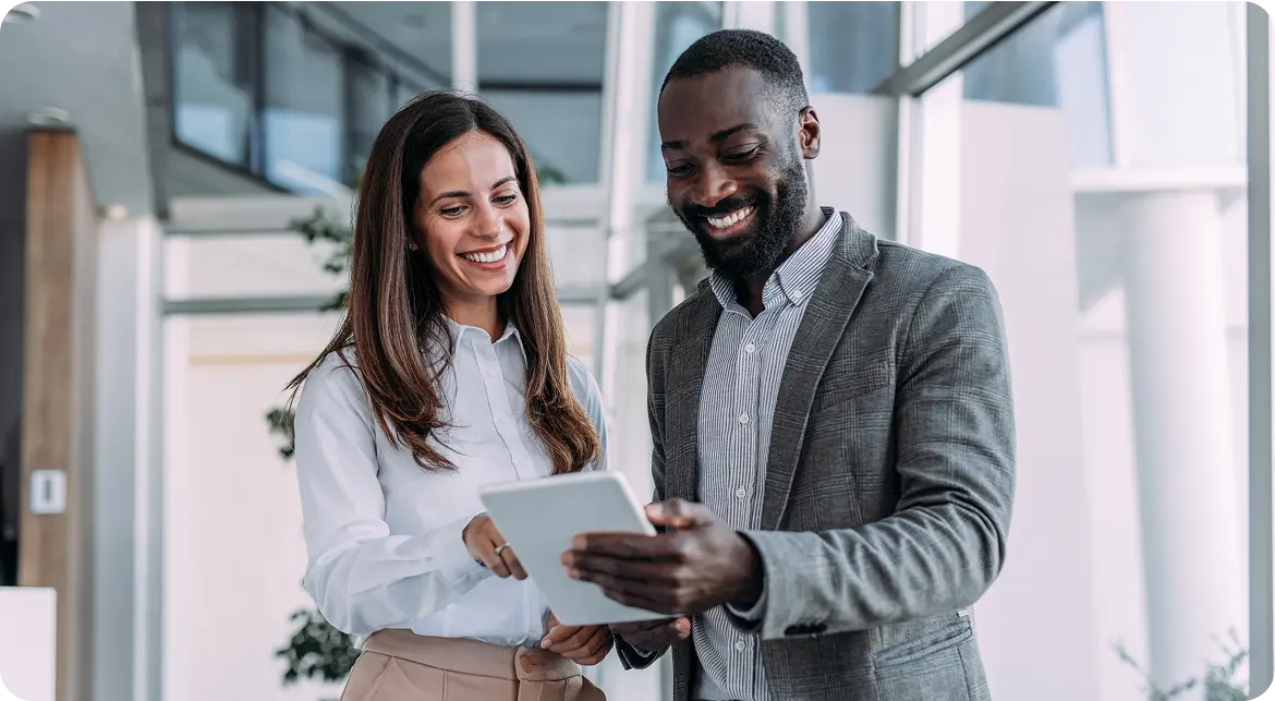 A woman with long brown hair and a man with a beard are smiling while interacting with a tablet in a modern office setting, standing near large windows.