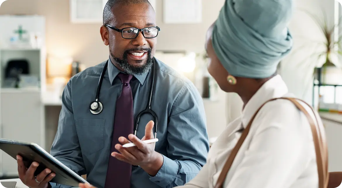 Hero-valenz-bluebook A doctor wearing glasses and a stethoscope around his neck is smiling and talking to a patient. The patient, facing away from the camera, is wearing a light-colored headscarf. They appear to be having a friendly conversation in a medical office setting.