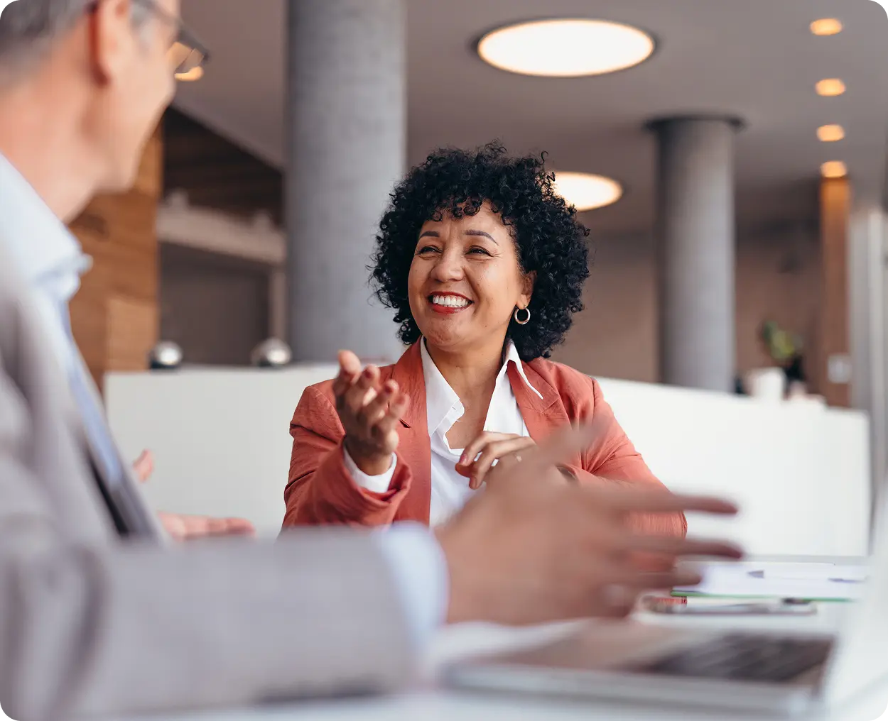 INN-Right A smiling woman with curly hair in a business suit is sitting at a table, engaged in a conversation with a blurred man in the foreground, suggesting a professional setting.