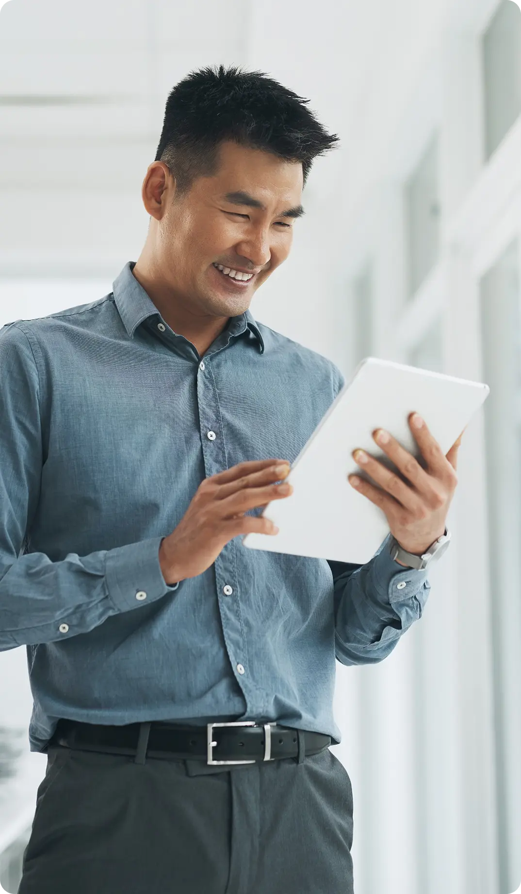 A man in a light blue button-down shirt smiles while using a white tablet in a bright, modern office environment.