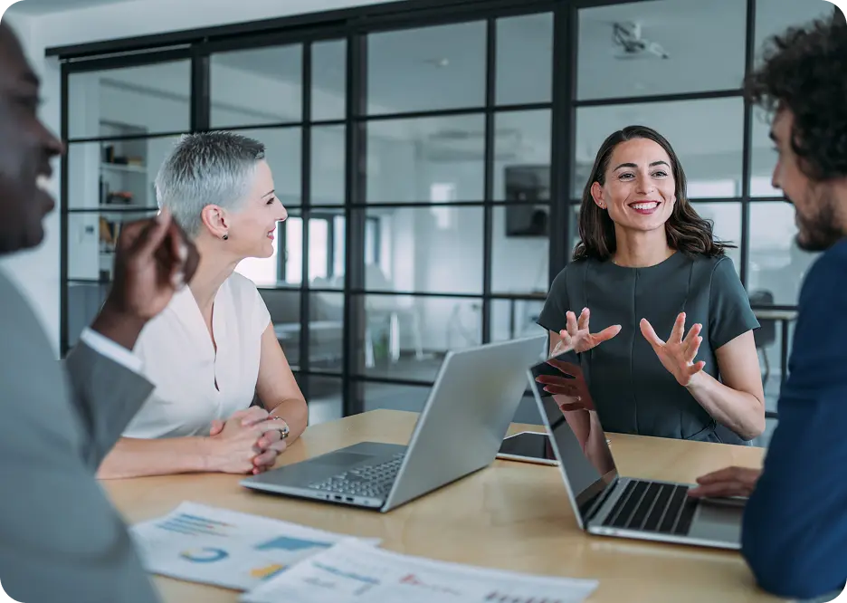 Payment-integreity-business-meeting A group of four professionals sitting around a table in an office, engaged in a discussion with laptops and papers in front of them. The woman in the center is speaking, gesturing with her hands, while the others listen attentively.
