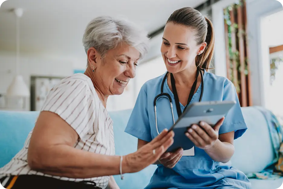 Plans-Performance-Right A healthcare professional in blue scrubs smiles while showing a tablet screen to an elderly woman with short gray hair, who is seated and looking pleased. The setting is a cozy living room with natural light.