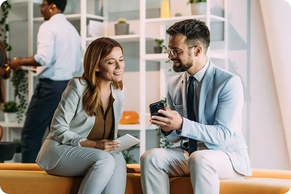 A woman in a light gray suit and a man in a light blue suit are sitting together on a bench in a modern office setting, smiling and looking at a smartphone, while a man in the background appears to be working with plants.