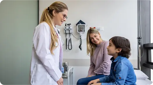 A female doctor speaking to a young boy sitting on an examination table, with a smiling woman, likely his mother, sitting nearby in a medical office.