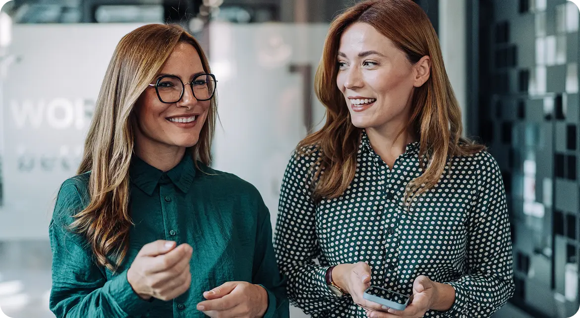 Two women smiling and conversing in an office setting, one holding a smartphone, both wearing patterned shirts.