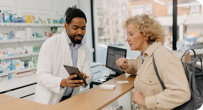 right-bluebook-rx A pharmacist showing a tablet screen to a customer at the pharmacy counter, with shelves stocked with medications in the background.
